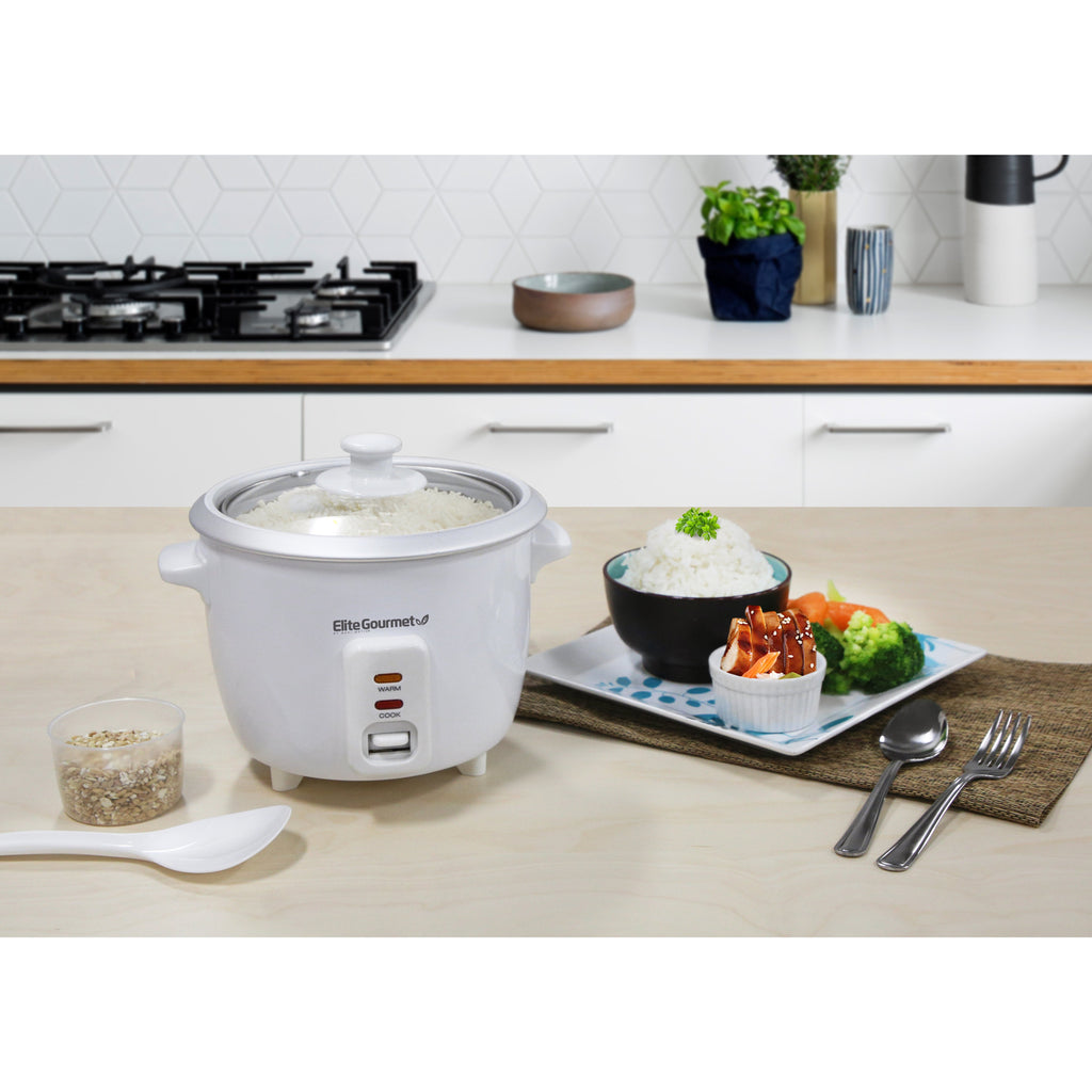 Rice cooker display on kitchen counter with cooked white rice inside pot.  Next to a plate of prepared food, rice and vegetables.