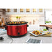 The metallic red slow cooker is displayed on kitchen counter next to serving dishes and food.