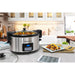 The stainless slow cooker is displayed on kitchen counter next to serving dishes and food.