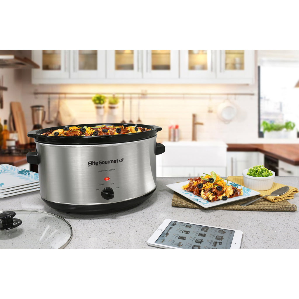 The stainless slow cooker is displayed on kitchen counter next to serving dishes and food.